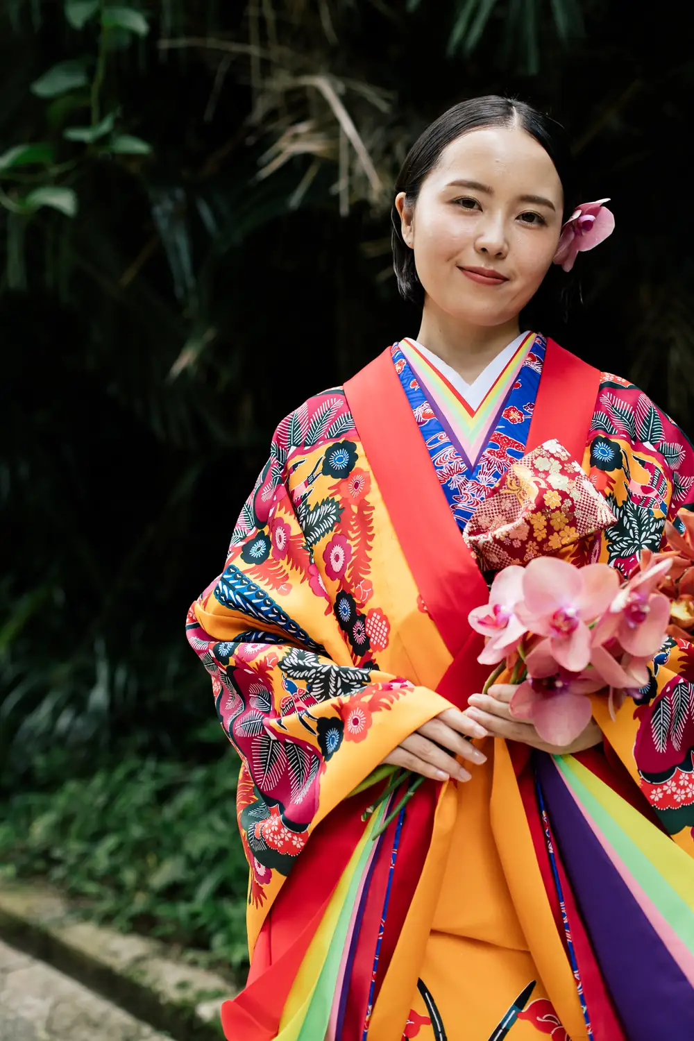 緑を背に、花を手に微笑む琉装の女性