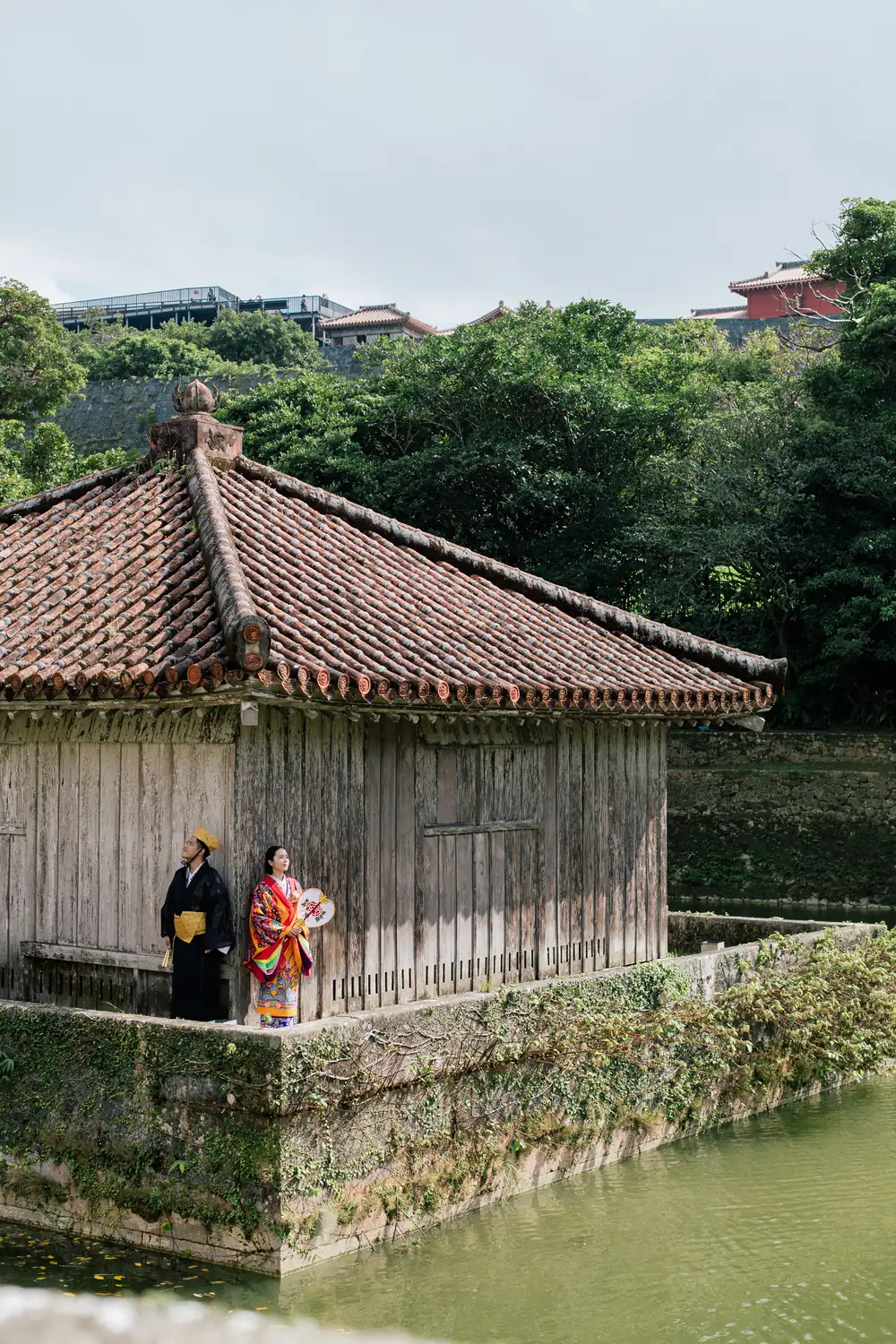 池のほとりの茅葺きの建物の前に立つふたり